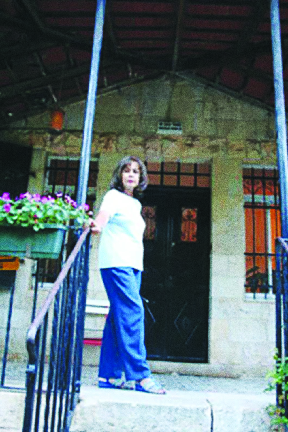Ghada Karmi standing by the front door of her childhood home in Jerusalem’s Qatamon neighborhood in 2005. She wears a t-shirt, blue pants, and sandals. Her hand is resting on the handrail by the stair on the front porch.